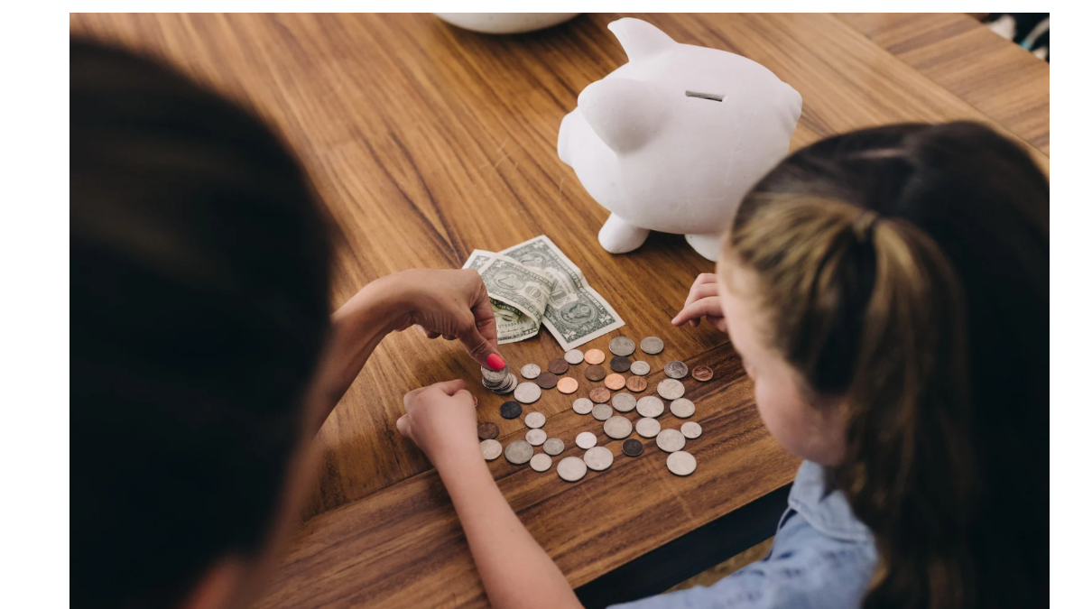 子どもと一緒にお金を数えながら、楽しそうに学び合う親子の手元。白いブタの貯金箱と紙幣・硬貨が机に並んでいる。